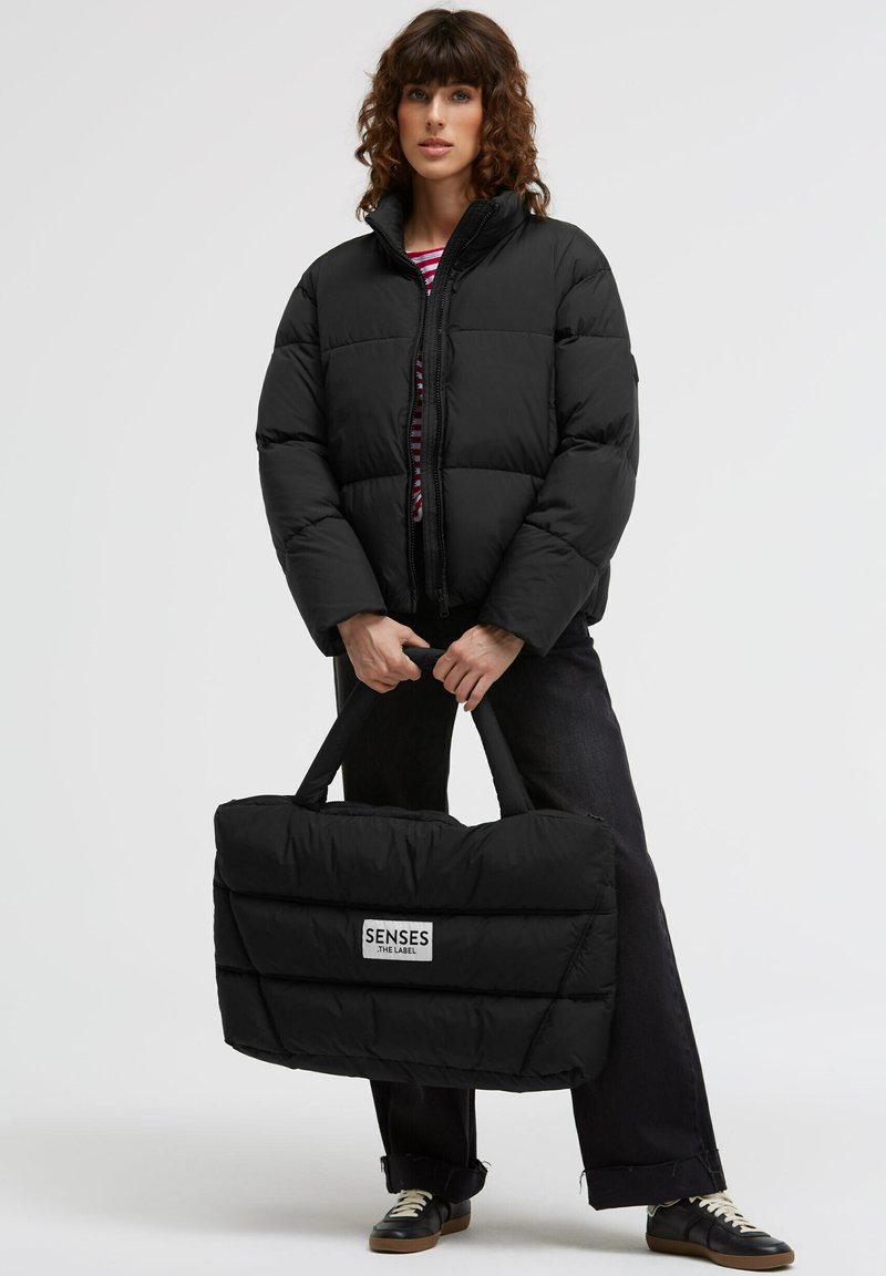 Schwarze Steppjacke mit hohem Kragen und Reißverschluss. Das Modell hält eine passende schwarze gesteppte Tote mit Logo. Dunkle Jeans und Schuhe.