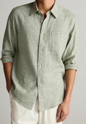 Homme portant une chemise en lin texturée vert clair à boutons avec manches retroussées et pantalon blanc, debout avec une main dans la poche.