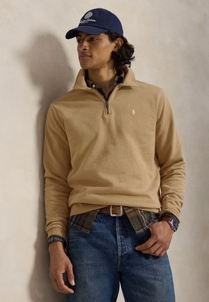 Ung mand iført en beige pullover med kvartlynlås, ternet skjorte, blå jeans, navy kasket og armbåndsur, stående mod en neutral baggrund.