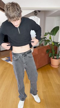 Fekete kötött pulóver cipzáras gallérral, szürke farmernadrággal és fehér sportcipővel. Az egyén a pulóvert felemeli, hogy megmutassa a hasát.