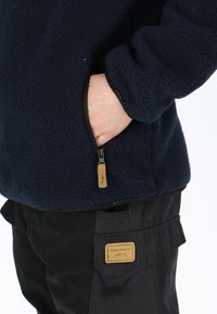 Giacca in pile navy con tasca con zip, caratterizzata da un tessuto testurizzato e polsini elasticizzati. Abbinata a pantaloni neri con una patch del logo.