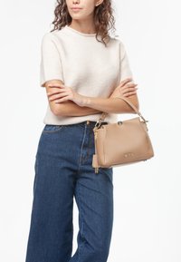 Borsa in pelle beige con accessori dorati, forma strutturata e dettagli con zip. Il modello indossa un maglione chiaro e jeans larghi scuri.
