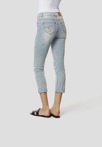 Világoskék farmer capri nadrág felhajtott szegéllyel, hátul díszítő varrással ellátott zsebekkel, fekete papucs szandálokkal viselve.