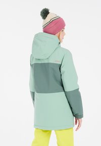 Chaqueta de invierno verde claro con panel horizontal verde oscuro, que presenta capucha y puños elásticos. Acompañada de un gorro de punto rosa con pompon.