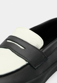 Schwarz-weiße Lederloafers mit glatter Textur. Sie verfügen über einen auffälligen schwarzen Riemen und ein Ausschnittdetail im Obermaterial.