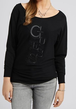 Kobieta ubrana w czarną bluzkę z długim rękawem z dużym pionowym logo „GUESS” i małymi ozdobami, w połączeniu z jasnoszarymi jeansami i srebrnym naszyjnikiem.