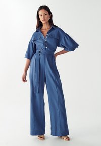 Kvinna står i en blå jumpsuit med bälte, vida ben och puffärmar i armbågslängd, bär beige klacksandaler.