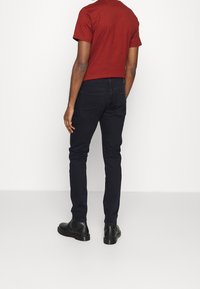 Mörkblå slim-fit jeans i kombination med en åtsittande röd t-shirt, visad bakifrån, med svarta ankbottnar. Slät tyg utan synliga mönster.