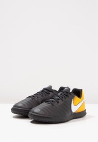 Botas de fútbol Nike en negro y amarillo con parte superior texturizada, cordones y un logo de swoosh blanco en el lateral; punta redondeada y suela de goma.