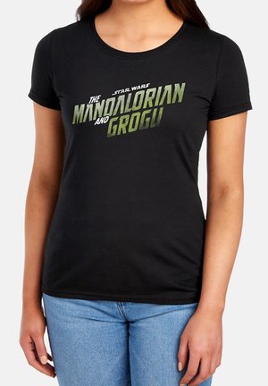 Frau trägt ein schwarzes T-Shirt mit grünem und weißem Text, auf dem "Star Wars The Mandalorian and Grogu" steht, kombiniert mit blauen Jeans.