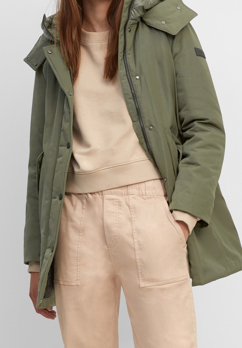 Parka verde oliva, resistente all'acqua, con chiusura a zip e bottoni, grande cappuccio e tasche frontali. Indossata sopra una felpa beige e pantaloni.