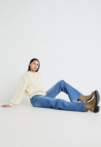 Maglione a maglia crema con motivo testurizzato, jeans a campana azzurri chiari e stivaletti marroni con tacco nero spesso, seduto su un pavimento bianco.