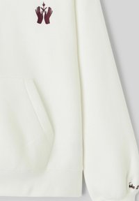 Sweat-shirt blanc avec poche kangourou, mains brodées en rouge foncé tenant une petite plante, et texte partiel sur le bord de la manche.