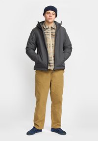 Chaqueta gris acolchada con capucha, camisa estampada en beige y marrón debajo, pantalones color tostado y zapatillas deportivas azules. Contra un fondo blanco.
