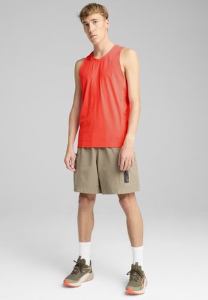 Haut de sport sans manches rouge, en matériau léger, associé à un short beige avec poches. Le modèle porte des baskets vertes et des chaussettes blanches.