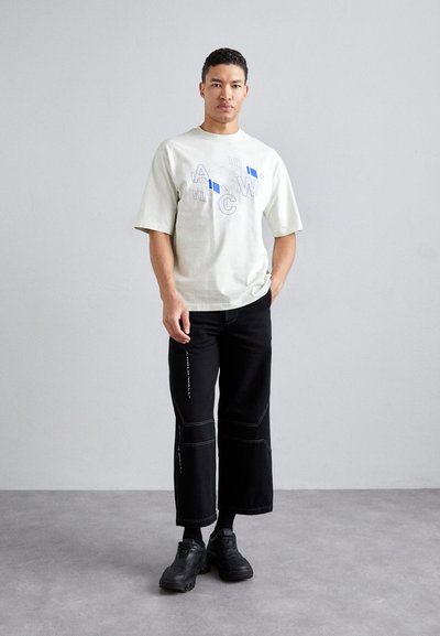 Lys beige oversized t-shirt med blåt grafisk design, parret med sorte brede bukser med hvid syning og sorte sko.