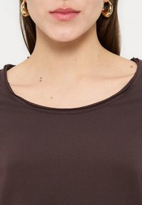 Γυναίκα που φορά χρυσούς κρίκους ως σκουλαρίκια και σκούρο καφέ μπλουζάκι με στρογγυλή λαιμόκοψη, εμφανίζονται τα χείλη, ο λαιμός και η περιοχή της κλείδας.