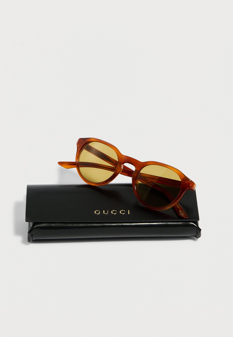Brown-tinted round sunglasses resting on a black Gucci case with gold lettering, shown on a plain white background.