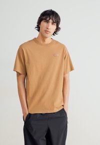T-Shirt mit kurzen Ärmeln und Rundhalsausschnitt in hellbraunem Baumwollstoff, mit einem kleinen Logo-Patch auf der linken Brust. Hergestellt aus einem weichen, leichtem Material.