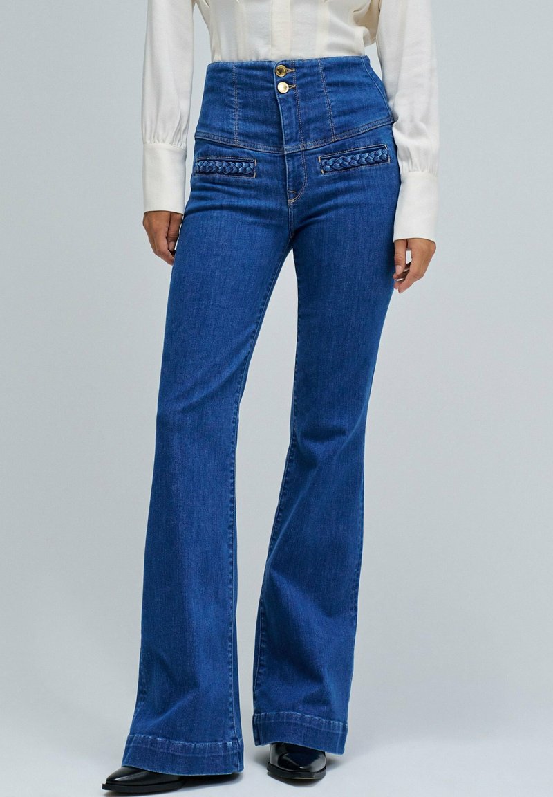 Pantalones vaqueros de cintura alta en azul con campana, con bolsillos trenzados en la parte frontal, cierre de dos botones y una textura suave. Footwear negro completa el conjunto.