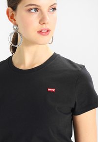 Svart bomull t-skjorte med rund hals, korte ermer, med et rødt Levi's-logo-patch på venstre bryst. Modellen har på seg store sølvringer som øreringer.
