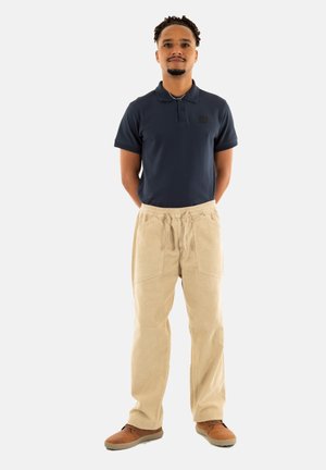 Marineblauwe polo, beige corduroy broek met elastische tailleband, zijzakken en bruine schoenen. Staande houding tegen een witte achtergrond.