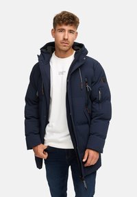 Mörkblå puffjacka med en texturerad design, dragkedjefickor och huva. Bärs över en vit skjorta och blå jeans. Har elastiska detaljer.