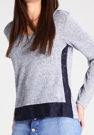 Femme portant un haut gris à manches longues avec des panneaux latéraux et un ourlet en dentelle noire, associé à une jupe en jean bleue à boutons.