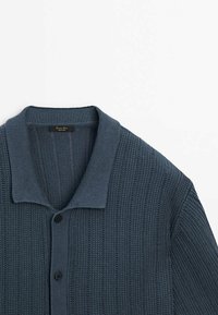 Cardigan en maille bleu foncé avec texture côtelée, fermeture par boutons et large col, soigneusement étalé à plat sur un fond blanc.