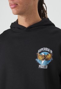 Čierna mikina s kapucňou, na prednej strane je grafika so zlatohnedým orlom na modrom kruhovom pozadí, text znie "SUPERBIA IN PROELIO", mäkký materiál.