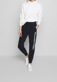 Persoon in een witte hoodie, zwarte Abercrombie & Fitch joggingsbroek met logo op het been, en witte sneakers, staande tegen een effen grijze achtergrond.