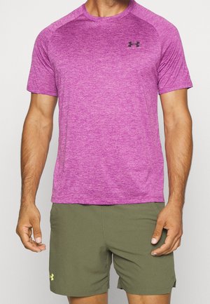 Homme portant un maillot de sport violet ajusté à manches courtes et un short vert olive Under Armour, debout devant un fond uni.