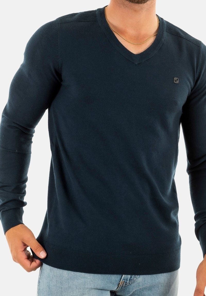 Homme portant un pull en V bleu foncé et un jean bleu clair, debout devant un fond uni.