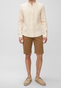 Uomo che indossa una camicia beige a maniche lunghe con bottoni, pantaloni corti marroni fino al ginocchio e scarpe beige con lacci, in piedi contro uno sfondo semplice.