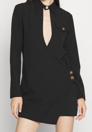 Sort langærmet slå-om kjole med dyb V-udskæring, guldknapper på krave, brystlomme og side, båret af en person med afslappede arme.