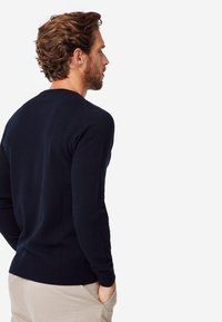 Pull en tricot bleu navy à manches longues et col rond ; texture lisse et coupe ajustée ; porté avec un pantalon de couleur claire.