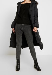 Manteau matelassé noir avec capuche, pull en maille noire texturé, jean skinny gris, et bottines noires brillantes à talons carrés.