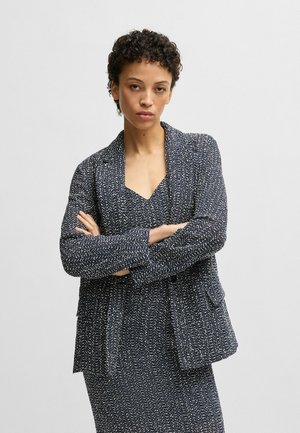 Navyfarbener Blazer mit Struktur in lockerer Passform, ausgestattet mit zwei Fronttaschen und einem Reverskragen, getragen über einem passenden Kleid.