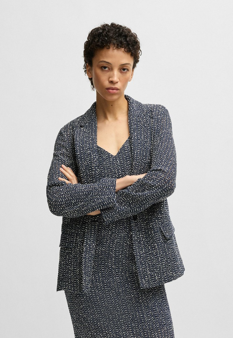Navy textured blazer with a relaxed fit, featuring two front pockets and a notched collar, worn over a coordinating dress.