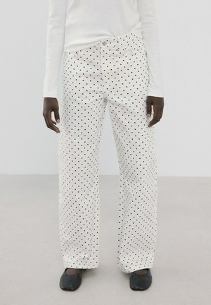 Personne portant un pantalon blanc à jambes larges avec des pois noirs, associé à un haut blanc à manches longues et des chaussures noires, debout sur un sol neutre.