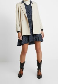 Blazer beige sopra un vestito blu navy con pois, caratterizzato da una gonna svasata. Stivali da cowboy neri e marroni con dettagli decorativi.