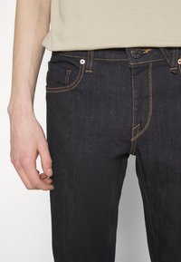 Homme portant un jean en denim bleu foncé avec des coutures orange et une chemise beige, la main reposant sur la cuisse gauche.