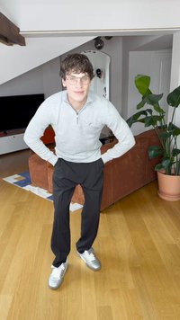 Lys grå genser med glidelås, svarte bukser og sølvfargede joggesko. Har strikket tekstur og smal passform. Innendørs miljø med plante og sofa.