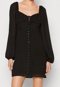Svart klänning i texturerad tyg, med fyrkantig halsringning, puffärmar, en frontknut och en central knappslå.