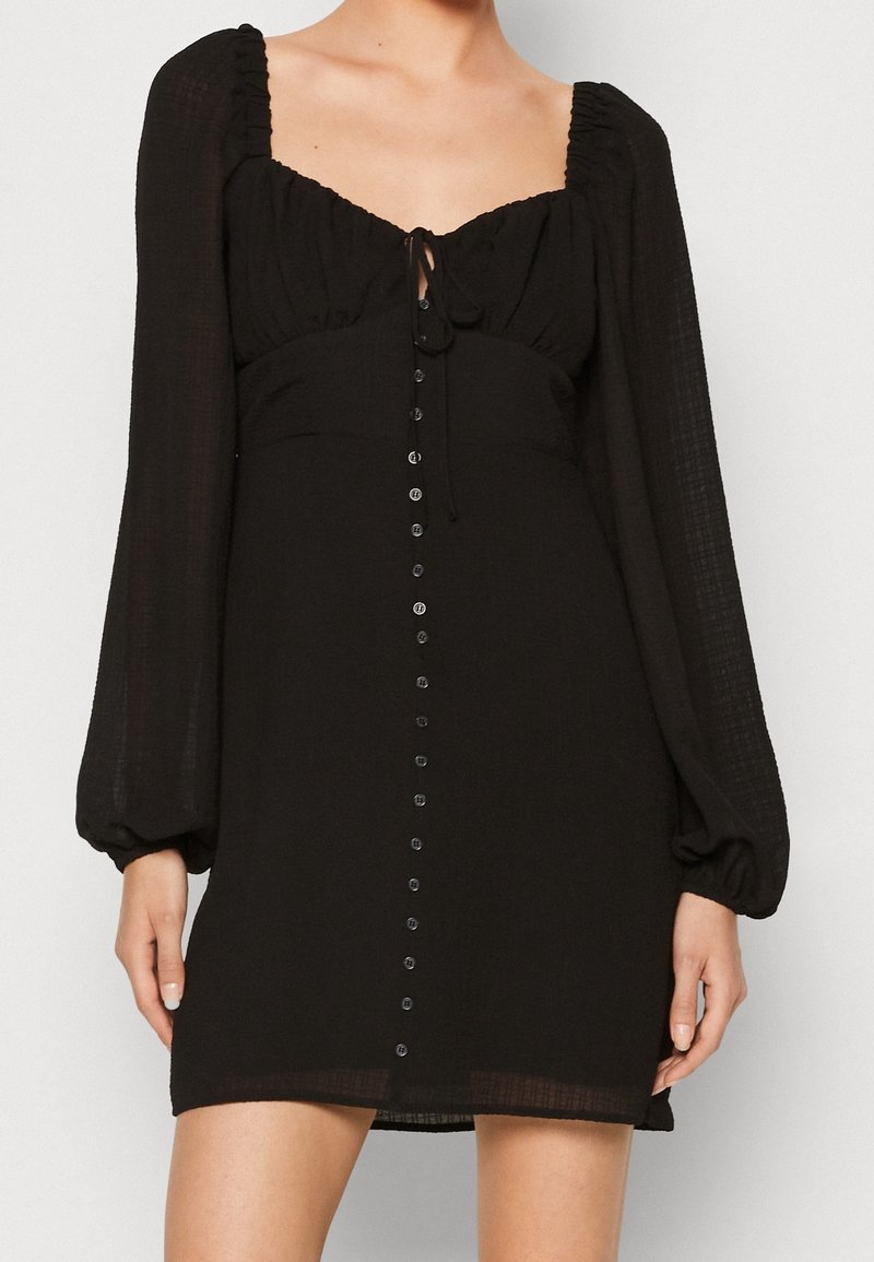 Svart klänning i texturerad tyg, med fyrkantig halsringning, puffärmar, en frontknut och en central knappslå.
