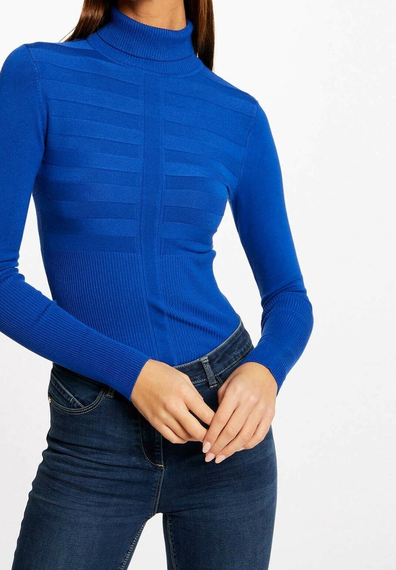 Femme portant un pull à col roulé bleu côtelé ajusté et un jean bleu foncé, les mains délicatement jointes au niveau de la taille.