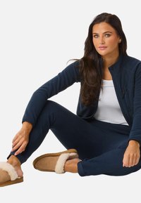 Femme aux longs cheveux foncés portant un survêtement bleu marine, un t-shirt blanc et des chaussons beiges, assise avec une jambe pliée et une main reposant sur sa cheville.