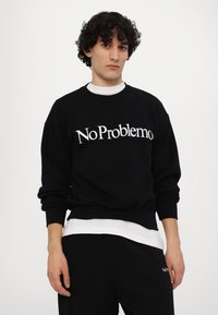 Schwarzer Sweatshirt mit fettem weißen "No Problemo"-Text, Rundhalsausschnitt, gerippte Bündchen und Saum, kombiniert mit einem weißen T-Shirt darunter, getragen mit schwarzen Hosen.