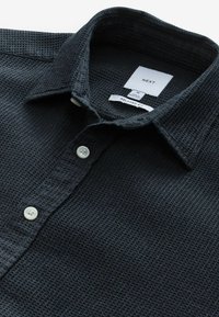 Chemise noire texturée, tissée en coton avec une coupe décontractée. Elle présente un col et trois boutons blancs le long de la patte de boutonnage.