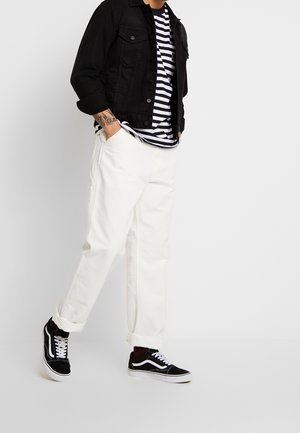 Schwarze Jeansjacke über einem gestreiften weißen und navyblauen Hemd, kombiniert mit weißen Hosen und schwarzen Sneakers mit weißen Akzenten.